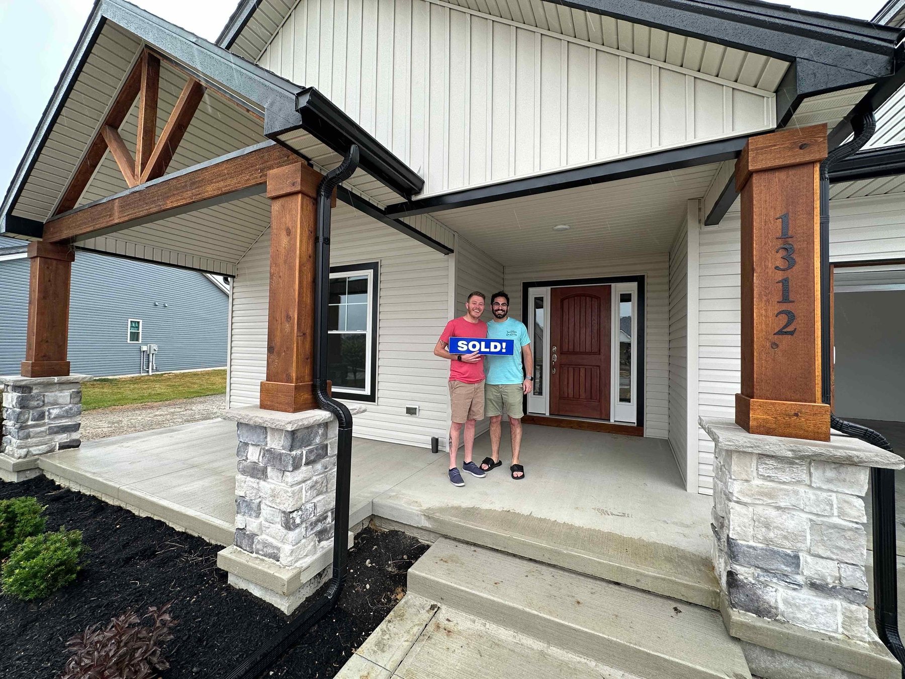 Justin and Brandon on the porch of their new house, holding a SOLD sign